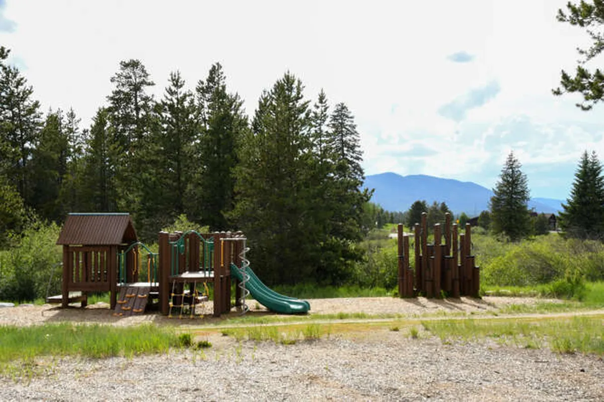 A vibrant outdoor playground set amidst lush green trees with striking green slides and a view of distant mountains. Children are seen playing, and the area invites families to gather and picnic while enjoying the fresh mountain air.