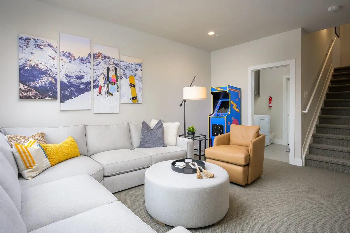 This image showcases a spacious game and lounge area featuring a large sectional sofa, an arcade game machine, a circular coffee table, and playful mountain landscape artwork. The TV and ample seating invite group relaxation and entertainment. The overall atmosphere is casual, lively, and well-suited for both family game nights and social gatherings.