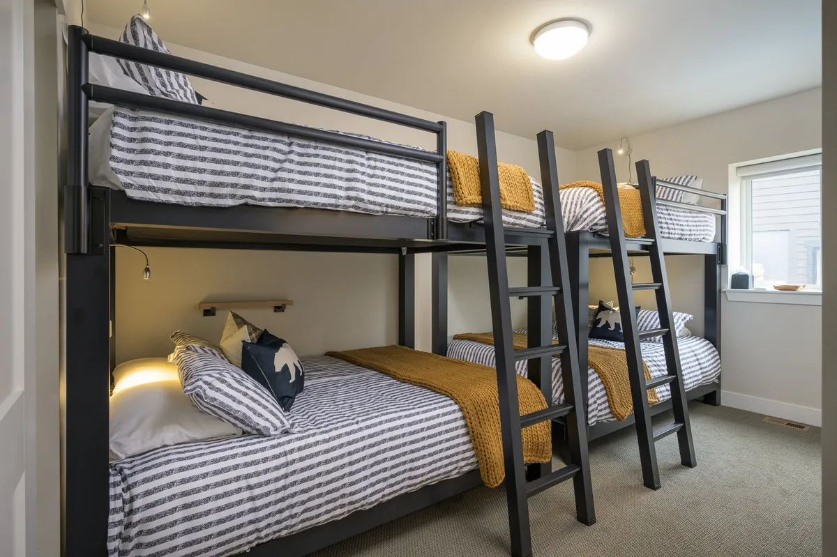 A playful and cozy bunkroom with two sets of queen bunk beds, striped bedding, cheerful yellow throws, and a large window allowing natural light to stream in. The layout provides ample sleeping space for kids or groups and a welcoming atmosphere for shared moments.