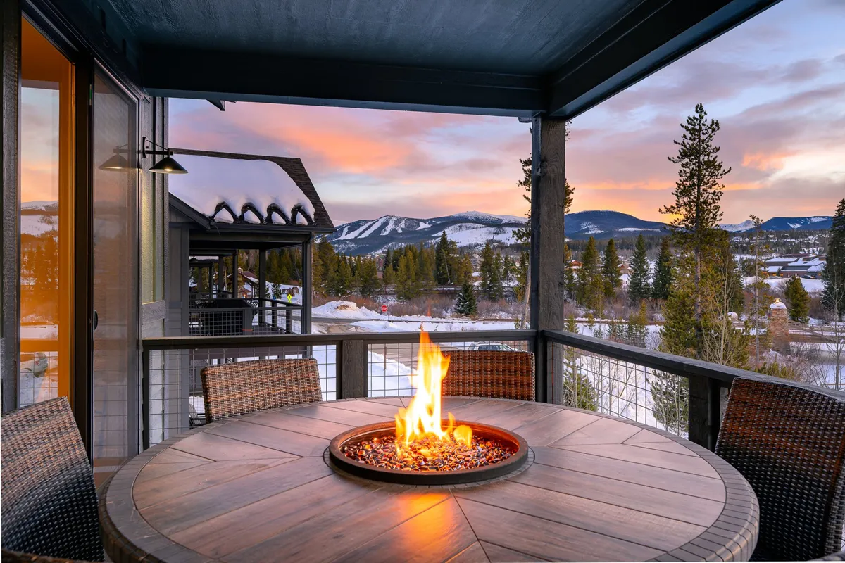 An inviting exterior dining area featuring a large table set near a modern fire pit, overlooking sweeping mountain views. Comfortable seating surrounds the fire table, and the atmosphere is cozy and intimate, perfect for small groups or couples. The sunset sky bathes the space in warm hues, while the mountain backdrop provides a sense of privacy and natural grandeur.