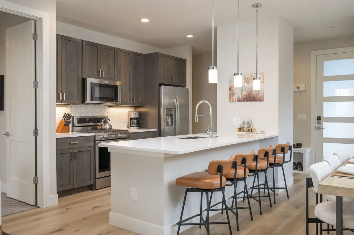 A contemporary kitchen scene featuring a spacious island with bar stools, stainless steel appliances, and stylish pendant lighting. The kitchen is designed for both cooking and socializing, with open sightlines to the living space and ample room for multiple guests.