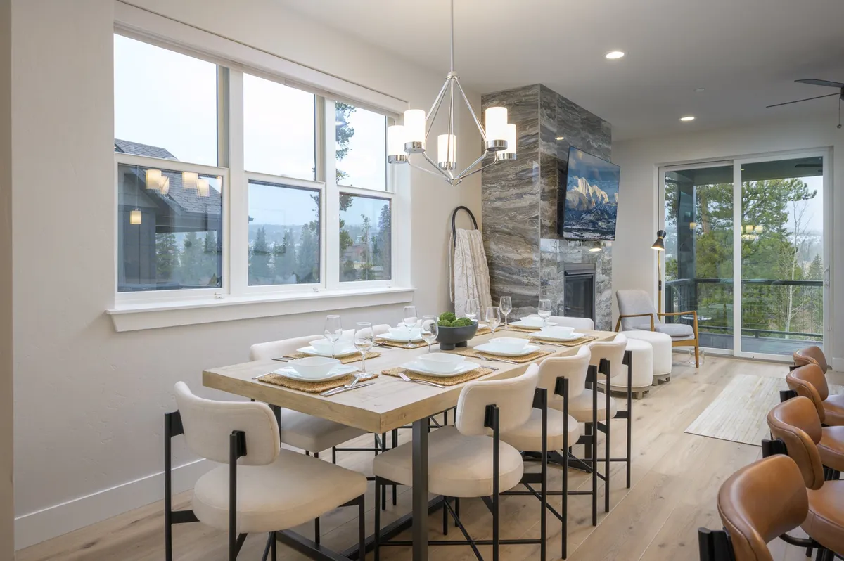 A warm, inviting dining area centered around a large table, complemented by a rustic stone fireplace and expansive windows that frame the surrounding mountain landscape. The setup is ideal for group dining and festive occasions.
