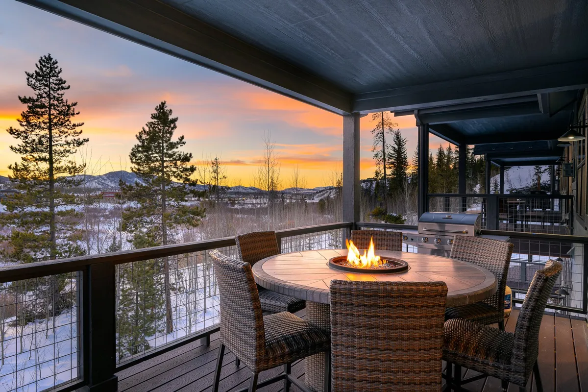 An outdoor scene with a fire pit and dining table set among trees, backed by distant mountains. The scene captures the golden hour, with the setting sun casting a romantic glow over the landscape. The arrangement is perfect for shared meals and gatherings in a tranquil, nature-filled environment.
