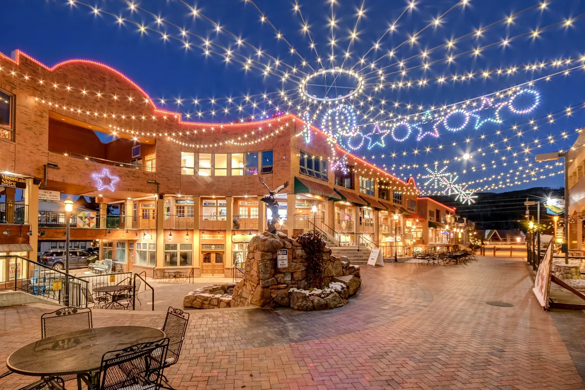 A lively downtown scene illuminated by string lights and decorative stars, with a stone fountain and inviting outdoor seating. The area is set up for dining, strolling, shopping, and socializing, evoking a festive atmosphere.