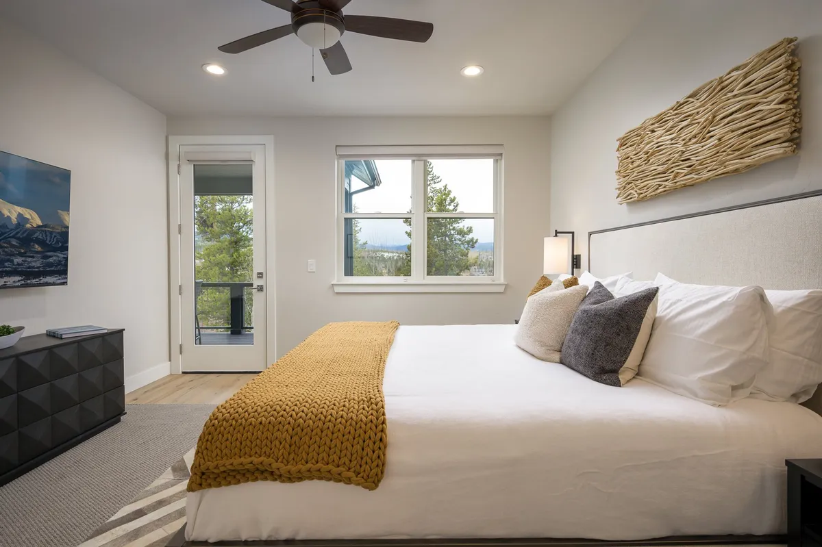 A serene king-sized bedroom featuring a plush bed adorned with decorative throws and pillows. Large windows frame scenic mountain vistas, and textured wall art adds a calming touch. The room exudes spa-like tranquility and offers a TV for relaxing entertainment.