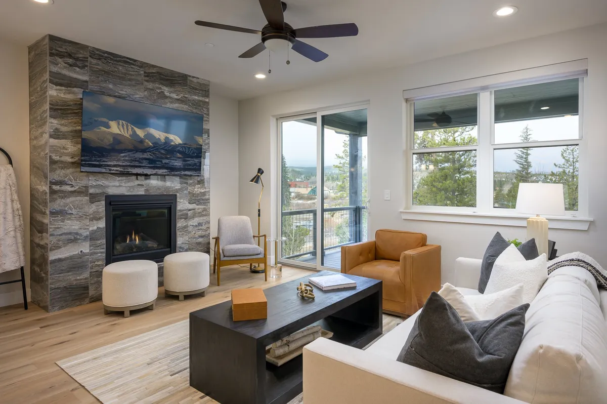 This scene highlights a comfortable living room with a plush sofa, coffee table, crackling fireplace, and oversized windows revealing an inspiring mountain panorama. The setup encourages relaxation and conversation, with a strong connection to the landscape outside.