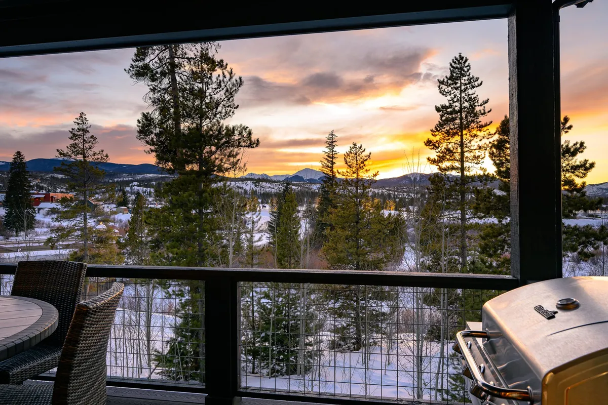 An inviting patio setup with furniture and a grill overlooks a tranquil sunset over the mountains. Distant trees and soft evening light create a peaceful ambiance, seamlessly blending outdoor living with the surrounding landscape.