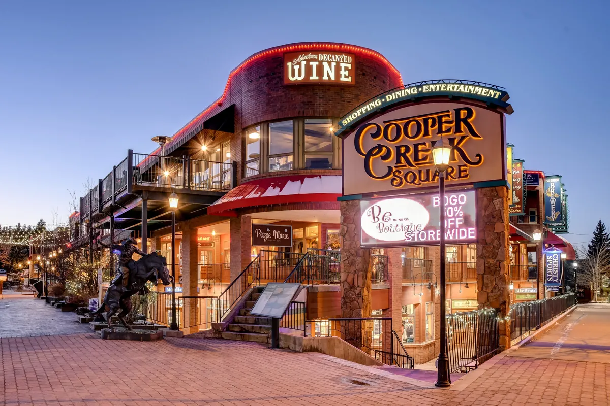 A festive downtown street scene featuring the Cooper Creek Square sign, boutique shops, decorative lighting, and a striking horse sculpture. The atmosphere is lively, hinting at shopping, dining, and entertainment options.