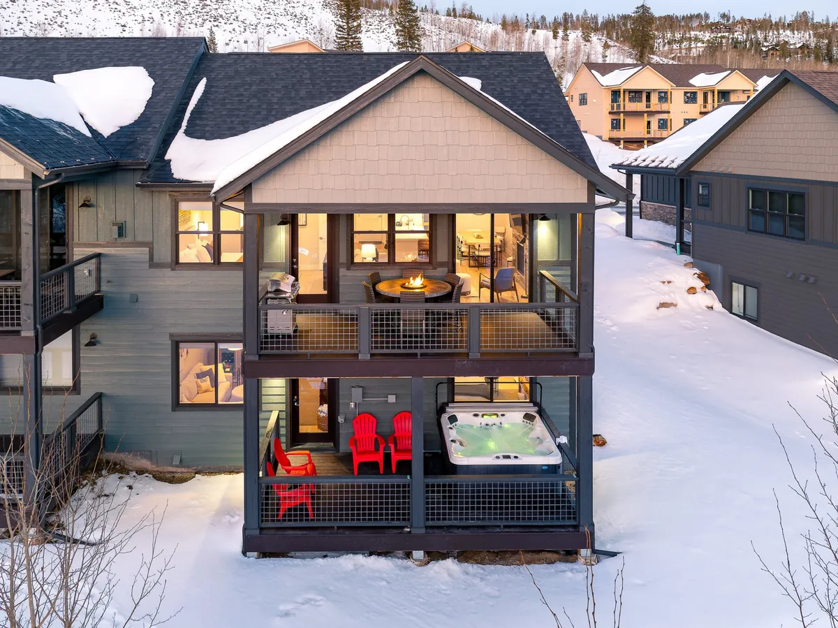 The image shows the inviting exterior of a luxury chalet, featuring a steaming hot tub surrounded by red Adirondack chairs, a cozy fire pit, and large windows that hint at warm, comfortable interiors. The setting is framed by scenic mountain views, creating an atmosphere perfect for relaxation and gathering with friends or family.