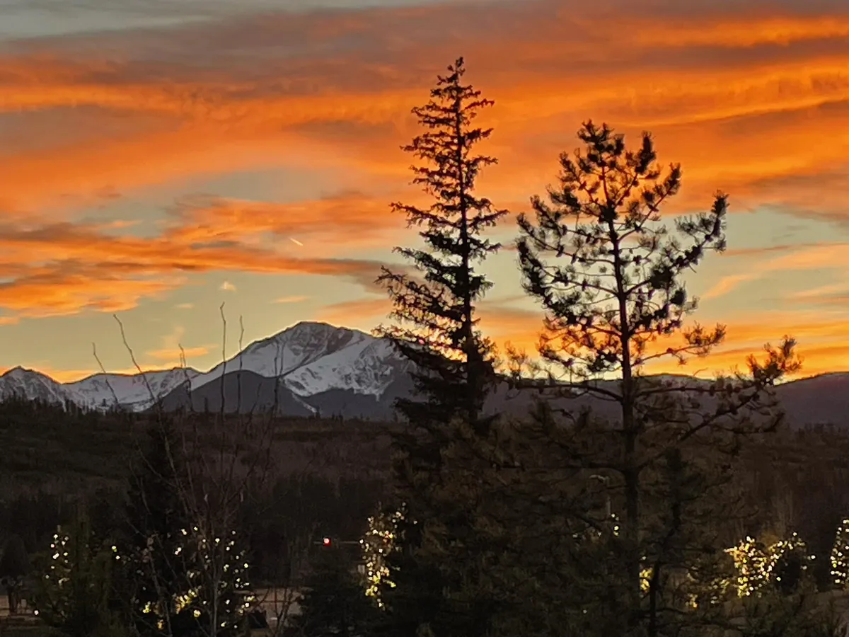 A sweeping mountain view captures snow-capped peaks under a vibrant sky, framed by silhouetted trees in the foreground. The scene evokes the serene, unspoiled natural beauty that surrounds the property, inviting exploration and connection with the Colorado environment.