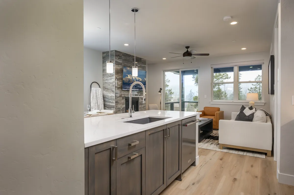 This image displays an open-concept living space with a kitchen island, living area, large windows, and a cozy fireplace. The design is modern yet comfortable, with abundant natural light flooding through the windows and comfortable seating arranged for social interaction. The mountain views visible through the windows create a seamless connection to the outdoors.