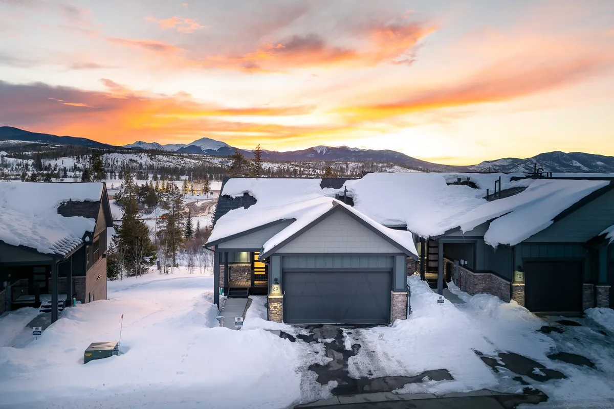 A tranquil winter exterior shot with snow-covered rooftops, mountains in the background, a vibrant sunset sky, and inviting front porches. The scene subtly suggests the presence of river views and direct access to nature right from the doorstep.